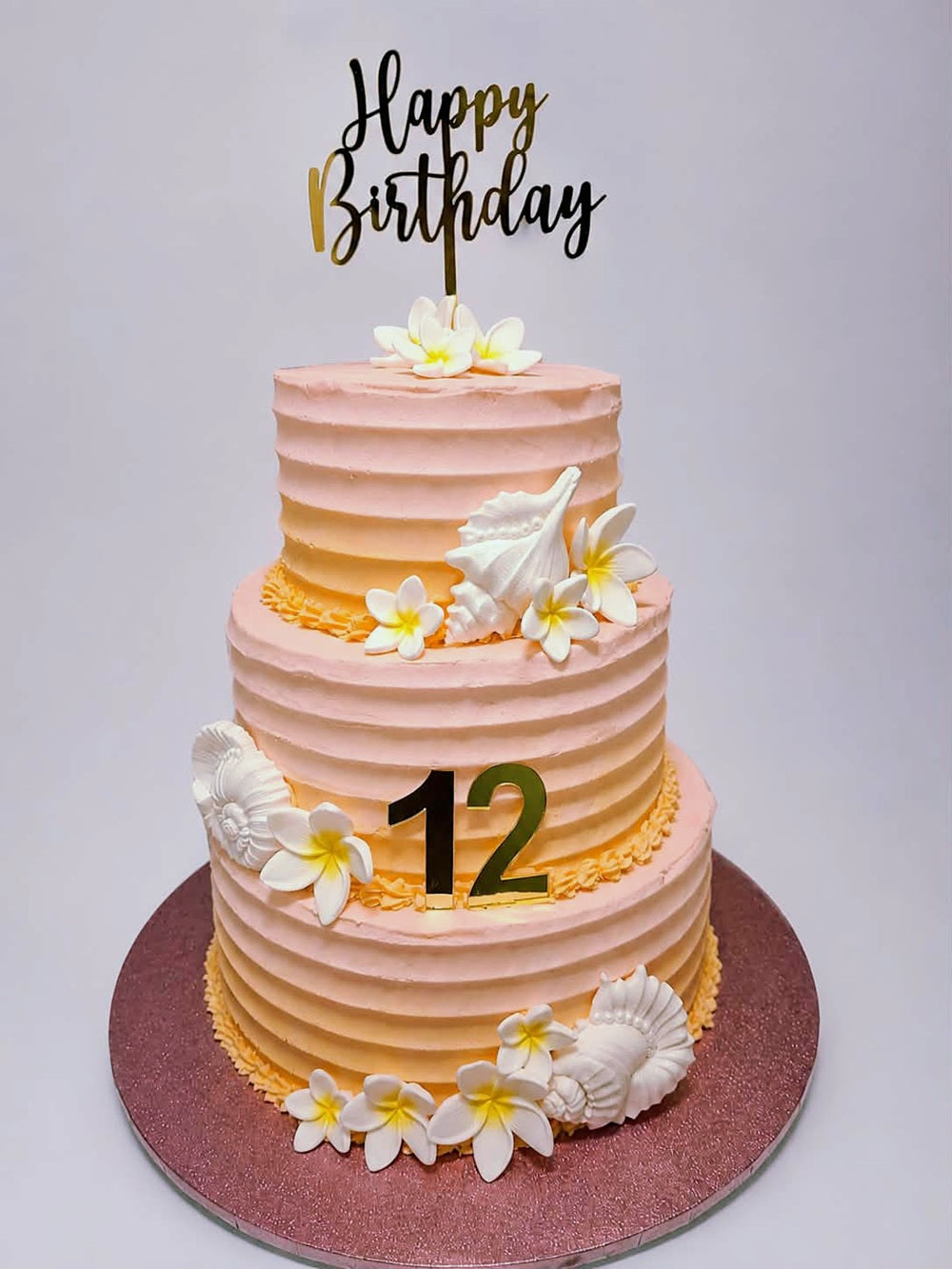 A three-tiered birthday cake with a pink and yellow gradient, decorated with white plumeria flowers, seashell decorations, and a gold 'Happy Birthday' topper. The cake is displayed on a glittery rose gold round board against a light gray background.