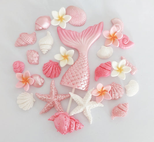 An assortment of fondant icing decorations including a mermaid tail, shells, and frangipani flowers, primarily in pink and white colors, arranged on a light background.