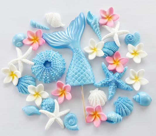 A collection of fondant icing decorations including a mermaid tail, shells, and frangipani flowers in blue, pink, and white colors, mounted on a white surface.