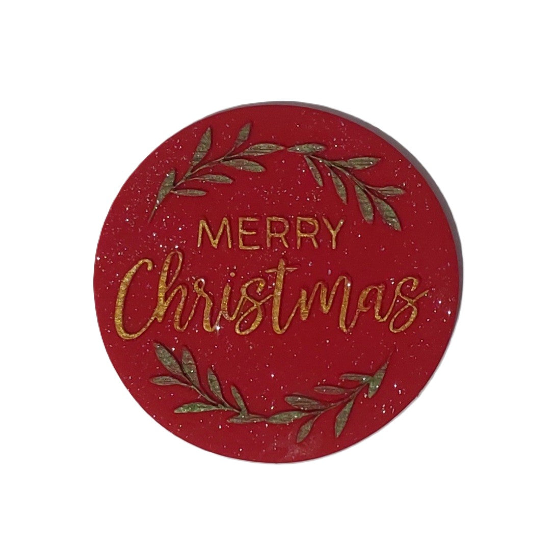 A round red decorative sign with gold glittery text reading 'MERRY Christmas' and green leafy branch accents, shown on a plain white background. Edible fondant decoration by Cake Candy NZ