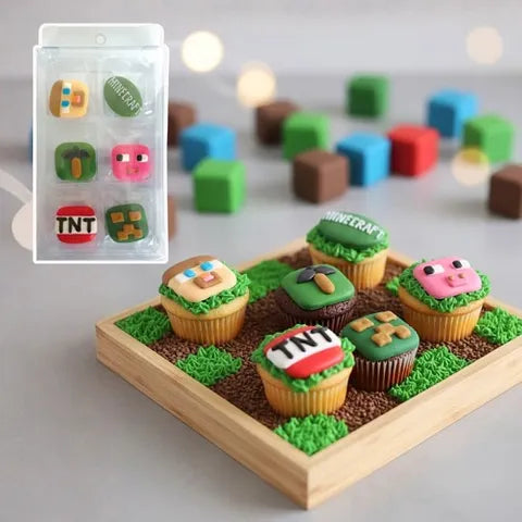 Minecraft-themed cupcake decorations and cupcakes displayed on a wooden tray, with a small inset showing the packaged set. The cupcakes are decorated with pixelated faces, grass, dirt, and block motifs including TNT, a creeper, a pig, and a Minecraft sign.
